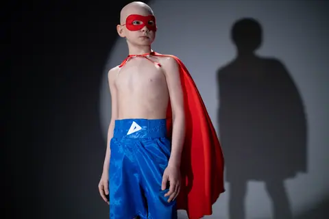Young boy standing strong in superhero attire with red cape and blue shorts, expressing bravery.