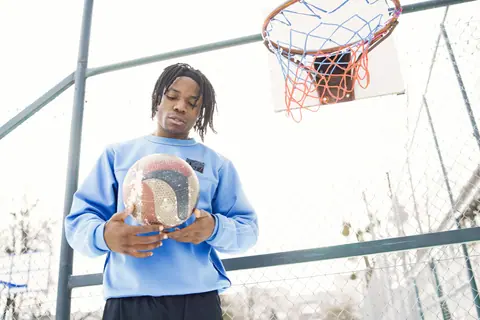 Young adult holding a basketball under an outdoor hoop in bright daylight.
