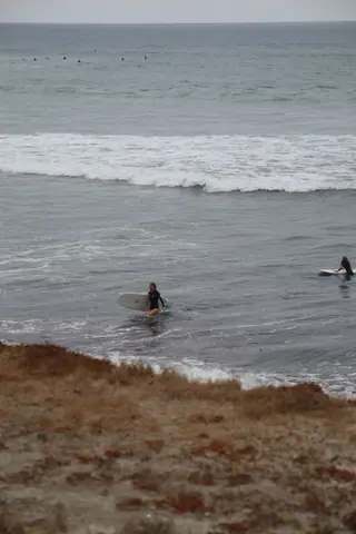 Surfers walking into the ocean with boards on a cloudy day at a rocky beach.