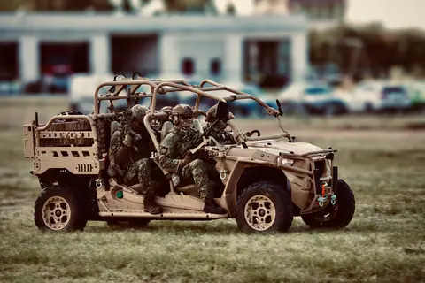 Military personnel wearing camouflage gear in off-road tactical vehicle on a grassy field.