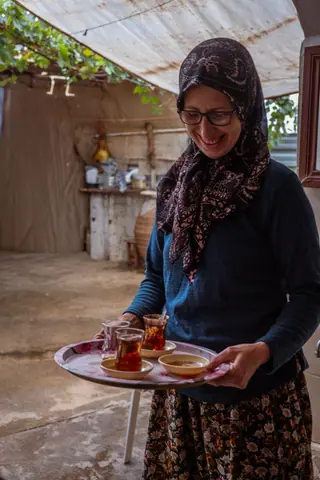 A woman with a headscarf serves tea on a tray in a rustic setting in Birgi, İzmir, Türkiye.