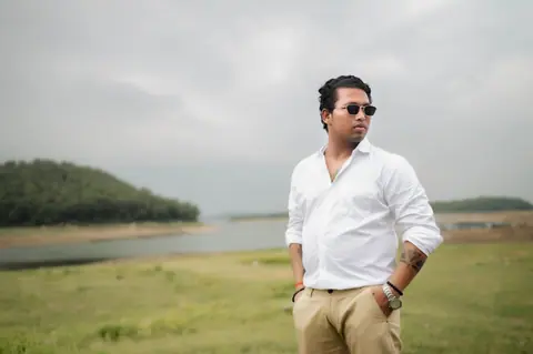Confident man in sunglasses and casual wear posing outdoors near a lake and grassy field.