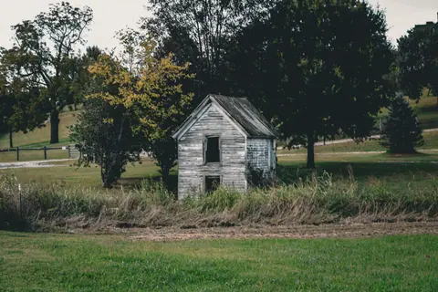 A weathered, abandoned house stands in a rural landscape amidst trees during autumn.