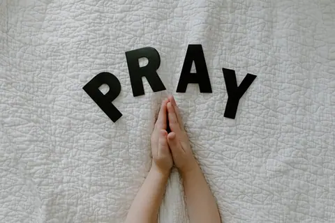 Close-up of hands in prayer on white quilted fabric with PRAY letters.