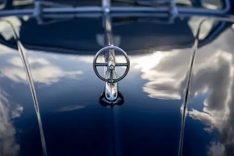 Classic vintage car hood ornament reflecting blue sky and clouds.