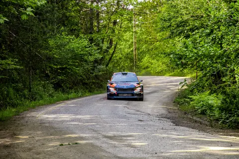 A rally car speeds through a lush forest path during a daytime race, capturing the thrill of motorsport in nature.