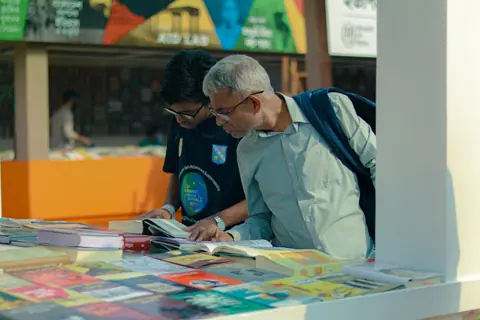 Two men explore books at an outdoor market table under a sunny day.
