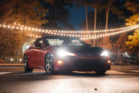 A red sports car with headlights on, parked under string lights at night.