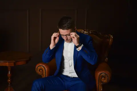 A well-dressed man in a blue suit adjusting his glasses while sitting in an armchair.