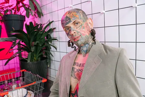 Portrait of a tattooed man in a blazer posing with plants indoors, modern aesthetic.