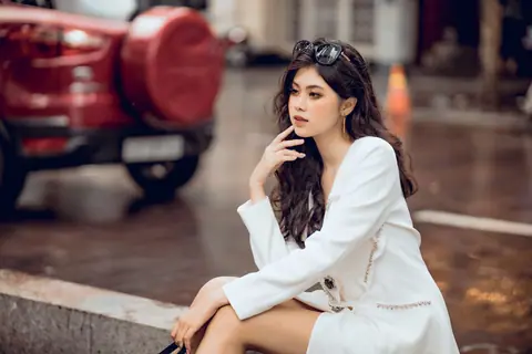 Fashionable woman in white sitting by street with blurred car background.