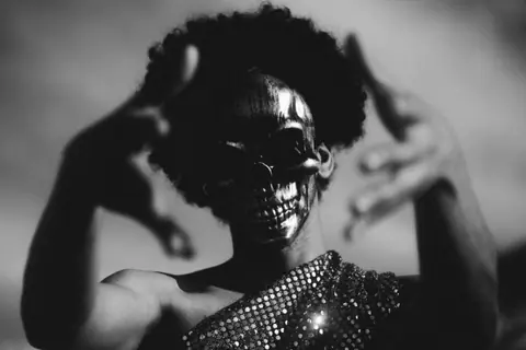 Black and white image of a person with afro hair wearing a skull mask and glittering attire, evoking mystery.