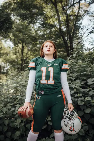 Redheaded female athlete holding football and helmet outdoors.