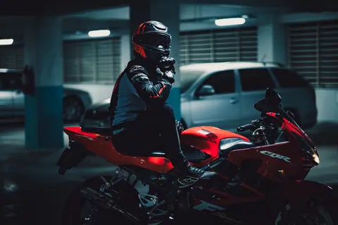 A motorcyclist with protective gear sitting on a red sport bike in a dimly lit parking garage.