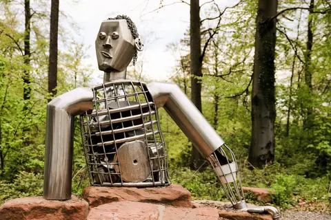 Artistic metallic sculpture of a robot figure in a serene forest environment.