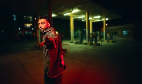 A stylish man posing at night by a gas station, showcasing fashion and dramatic lighting.