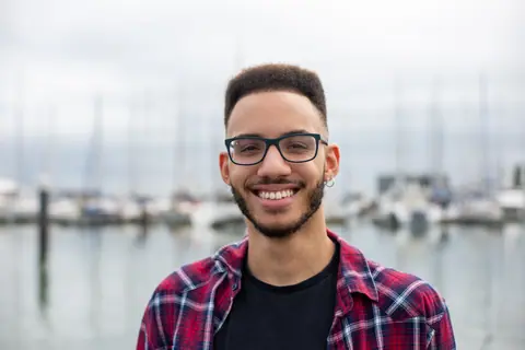 Cheerful young man with glasses and beard smiling by a marina in Portugal.