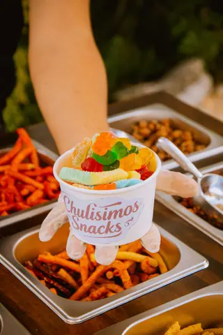 Assorted gummy candies in a snack cup offered by a vendor's hand wearing gloves.