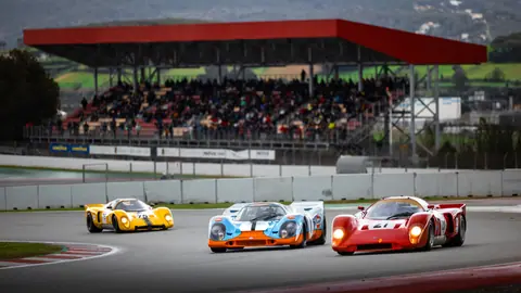 Three vintage race cars speed around a corner on a busy motor racing circuit.
