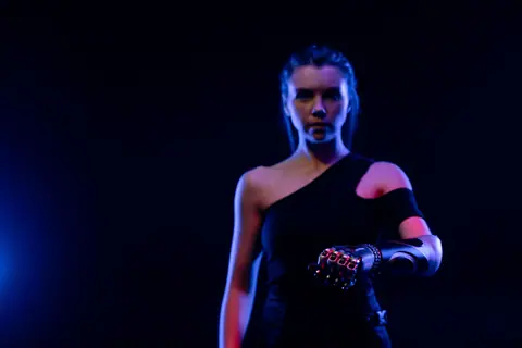 A dramatic studio portrait of a woman with a cutting-edge prosthetic arm under blue lighting.