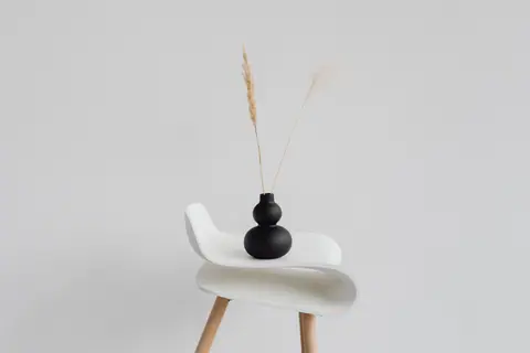 A sleek black vase with wheat ears on a modern white chair against a minimal background.