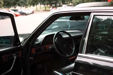 Close-up of an open car door showing the steering wheel and dashboard with a blurred parking lot background.