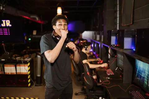 A young gamer excitedly posing in a dimly lit esports arena. Others playing in the background.