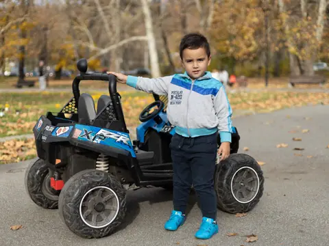 A young boy in a park standing beside a toy car, enjoying a sunny day outdoors.