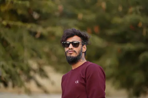 Confident man sporting sunglasses and a CK sweater with lush greenery backdrop in Mumbai.