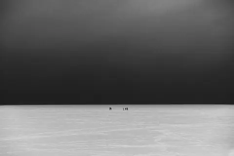 Minimalist black and white photo of vast snowy plains with distant figures.
