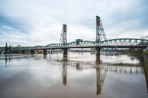 Steel drawbridge spans across a calm river, reflecting against cloudy sky.