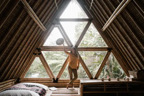 Woman standing in a wooden bamboo house with triangular windows, surrounded by tropical plants.