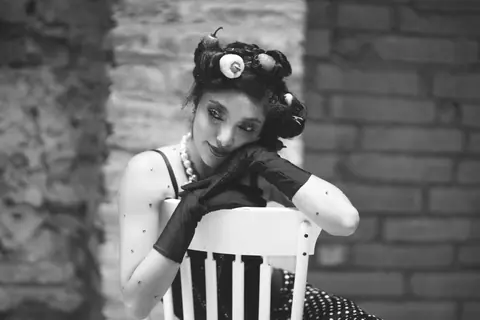 Black and white portrait of an elegant woman with pearls and gloves, sitting in a vintage setting.