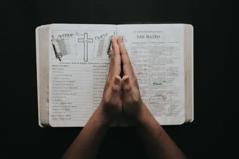 Hands clasped in prayer on an open Bible showing the book of Matthew, emphasizing faith and spirituality.