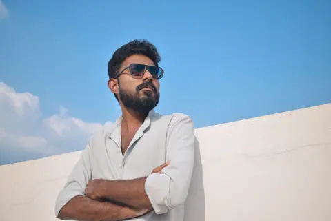 A stylish man in sunglasses stands confidently against a bright blue sky.