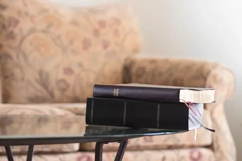 Close-up of a Holy Bible stacked on another book on a glass table with a patterned sofa in the background.