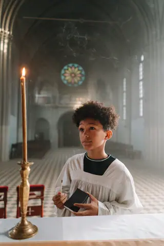 Young altar boy holding a bible near a candle in a serene church setting.