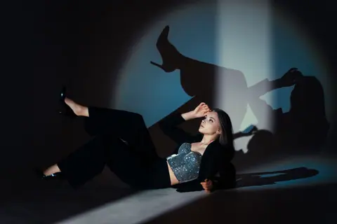 Stylish woman in shimmering top poses creatively indoors with bold shadows.