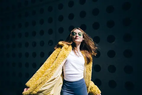 Confident woman wearing sunglasses and a yellow fur coat poses against a dotted wall in daylight.