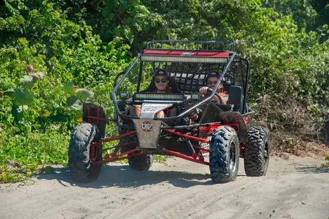 Two adults driving a dune buggy on a sandy trail through a lush green forest.