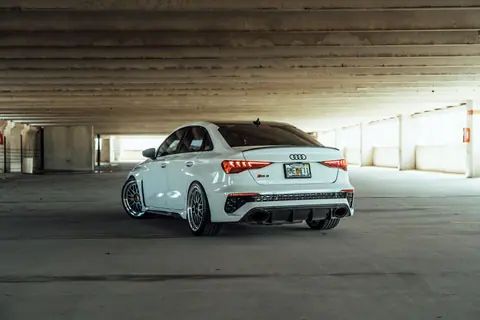 White Audi RS3 parked in a dimly lit, modern, urban parking garage. Rear view accentuates sporty design.