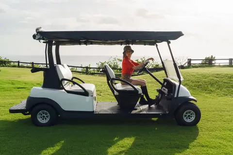 A woman driving a golf cart on a lush, sunlit golf course with ocean views.