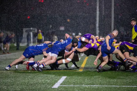 Dramatic rugby scrum at night in Manchester rain, showcasing teamwork and athleticism.