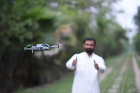 A man gives a thumbs-up while operating a drone in a forested pathway.