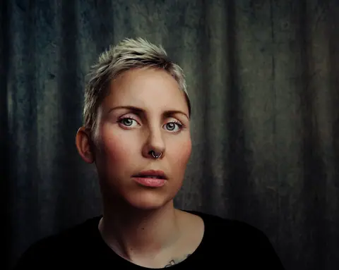 A striking portrait of a woman with short blonde hair, a nose piercing, and a contemplative expression.