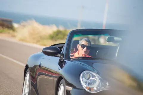 Elderly woman with short hair drives a convertible on a scenic road in Portugal.
