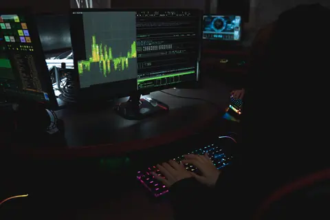 A cybersecurity expert in a dimly lit room is typing on a colorful keyboard with multiple screens displaying data.