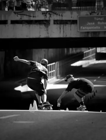 Two skateboarders performing tricks under a city bridge in a dynamic black and white capture.