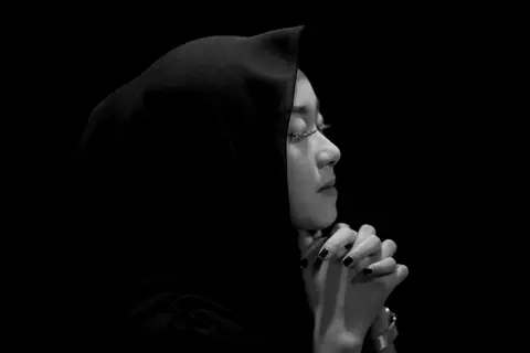 Profile of a woman in a hijab with hands clasped in prayer on a black background.