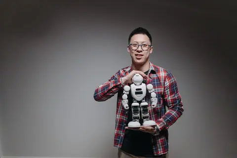 Asian man with eyeglasses holding a toy robot in a studio with a gray background.